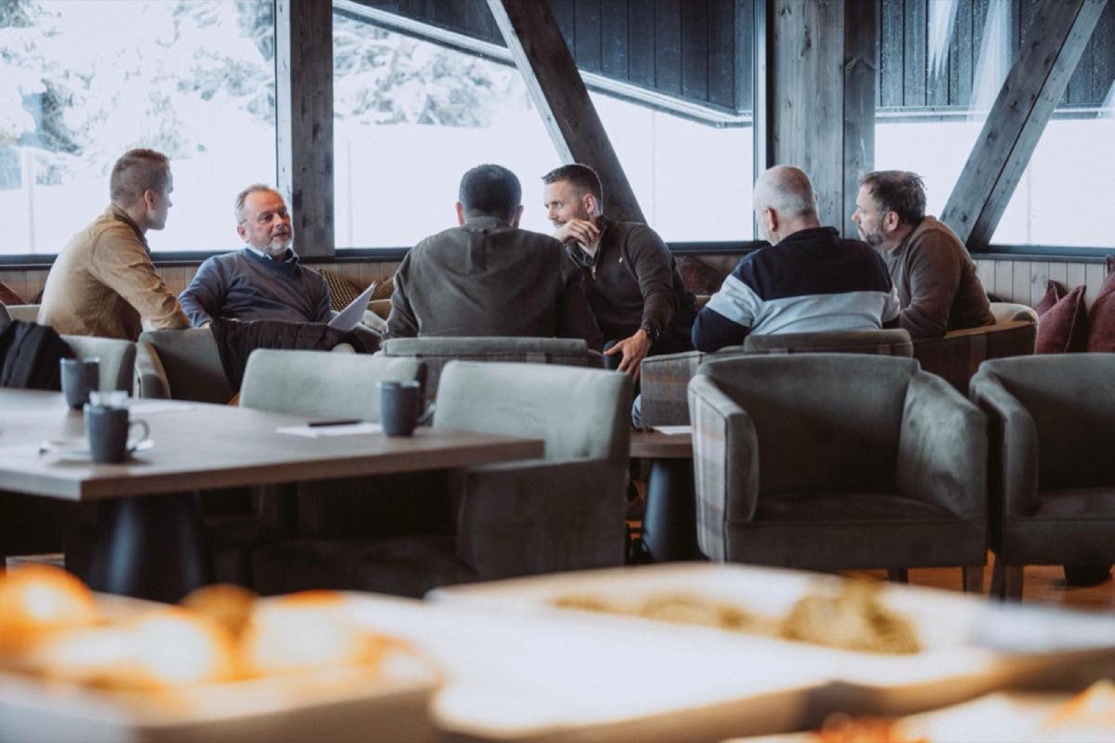 En gruppe menn i samtale rundt et bord i en lys fjellhytte-lounge, med snø utenfor vinduene